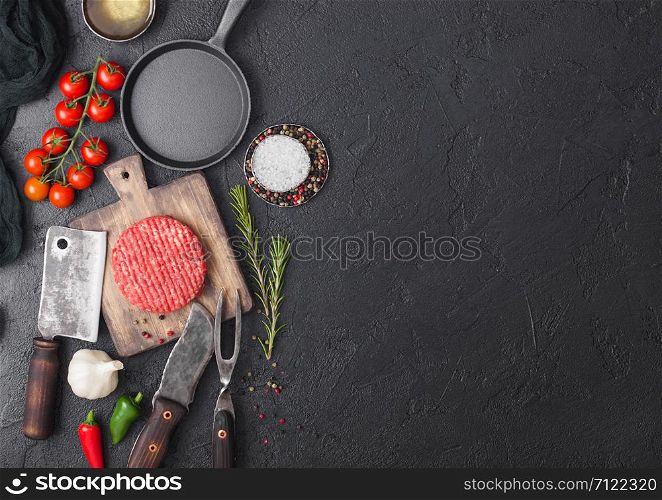 Fresh raw minced homemade farmers grill beef burgers on round chopping board and frying pan with spices and herbs and meat hatchet on black board. Space for text