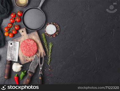Fresh raw minced homemade farmers grill beef burgers on round chopping board and frying pan with spices and herbs and meat hatchet on black board. Space for text