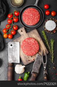 Fresh raw minced homemade farmers grill beef burgers on round chopping board and frying pan with spices and herbs and meat hatchet on black board.