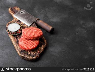Fresh raw minced homemade farmers grill beef burgers on vintage chopping board with spices and herbs and meat hatchet on black background.