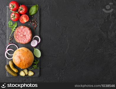 Fresh raw minced pepper beef burger on stone chopping board with buns onion and tomatoes on black background. Salty pickles and basil. Top view