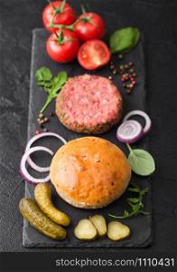 Fresh raw minced pepper beef burger on stone chopping board with buns onion and tomatoes on black background. Salty pickles and basil. Top view
