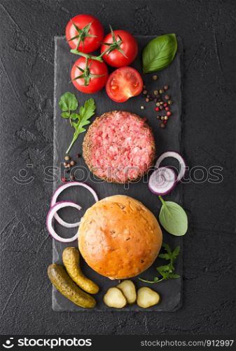 Fresh raw minced pepper beef burger on stone chopping board with buns onion and tomatoes on black background. Salty pickles and basil. Top view