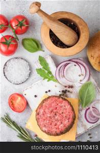Fresh raw minced pepper beef burger on vintage chopping board with buns onion and tomatoes on wooden background.Mortar with pestle with pickles and basil. Top view