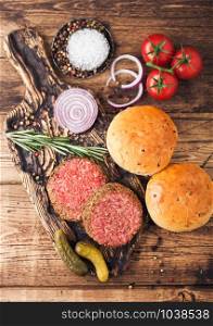Fresh raw minced pepper beef burgers on vintage chopping board with buns onion and tomatoes on wood background.