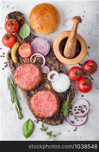 Fresh raw minced pepper beef burgers on vintage chopping board with buns onion and tomatoes on wooden background.Mortar with pestle with pickles and basil. Top view