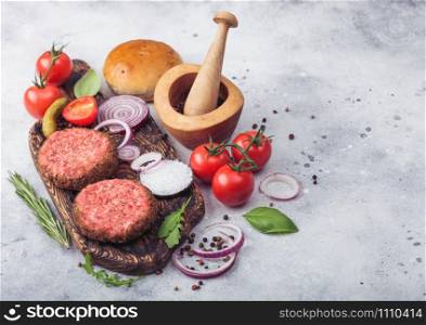 Fresh raw minced pepper beef burgers on vintage chopping board with buns onion and tomatoes on wooden background.Mortar with pestle with pickles and basil. Space for text