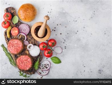 Fresh raw minced pepper beef burgers on vintage chopping board with buns onion and tomatoes on wooden background.Mortar with pestle with pickles and basil. Space for text