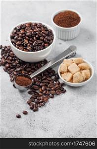 Fresh raw organic coffee beans in white bowl and powder on ligh table background with cane sugar and round steel scoop.