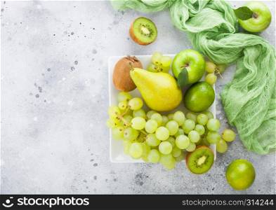 Fresh raw organic green toned fruit on white ceramic square plate on stone kitchen background. Pear and grapes with kiwi and lime and apples.