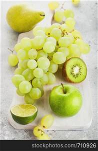 Fresh raw organic green toned fruit on white chopping board on stone kitchen table background. Pear and grapes with kiwi and lime and apple.