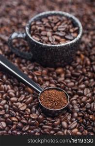 Fresh raw organic ground coffee powder in black steel scoop with black ceramic cup on top of coffee beans. Macro