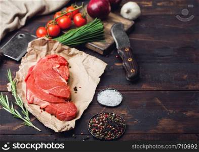 Fresh raw organic slice of braising steak fillet on butchers paper with fork and knife on dark wooden background. Red onion, tomatoes with salt and pepper and herbs.