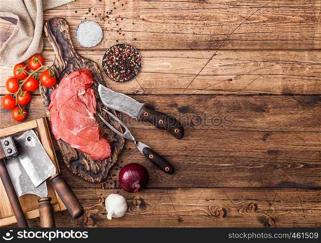 Fresh raw organic slice of braising steak fillet on chopping board with fork and knife on wooden background. Red onion, tomatoes with salt and pepper and kitchen towel.