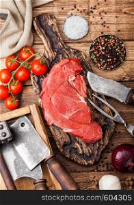 Fresh raw organic slice of braising steak fillet on chopping board with fork and knife on wooden background. Red onion, tomatoes with salt and pepper and kitchen towel.
