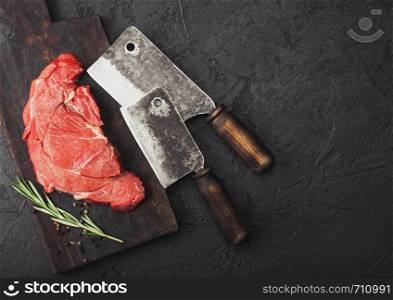 Fresh raw organic slice of braising steak fillet on chopping board with meat hatchets on black background.