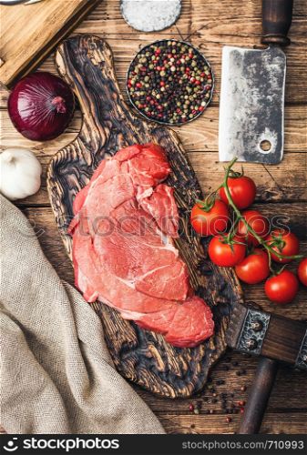 Fresh raw organic slice of braising steak fillet on chopping board with meat hatchet on wooden background. Red onion, tomatoes with salt and pepper and kitchen towel.