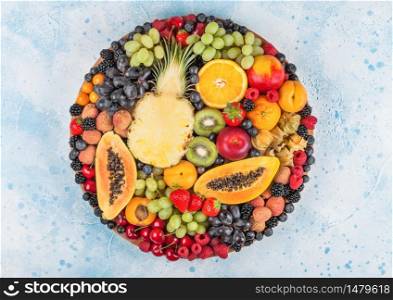 Fresh raw organic summer berries and exotic fruits in round large tray on blue kitchen background. Papaya, grapes, nectarine, orange, raspberry, kiwi, strawberry, lychees, cherry. Top view
