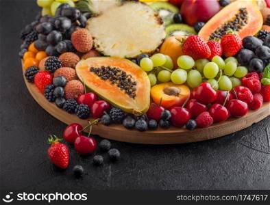 Fresh raw organic summer berries and exotic fruits in round large tray on black kitchen background. Papaya, grapes, nectarine, orange, raspberry, kiwi, strawberry, lychees, cherry. Top view