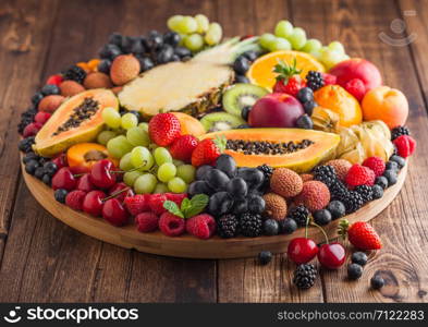 Fresh raw organic summer berries and exotic fruits in round large tray on wooden kitchen background. Papaya, grapes, nectarine, orange, raspberry, kiwi, strawberry, lychees, cherry.Top view