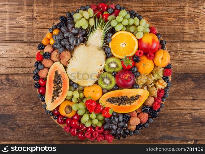 Fresh raw organic summer berries and exotic fruits in round large tray on wooden kitchen background. Papaya, grapes, nectarine, orange, raspberry, kiwi, strawberry, lychees, cherry.Top view