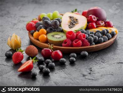 Fresh raw organic summer berries and exotic fruits in round wooden plate on black kitchen background. Papaya, grapes, nectarine, orange, raspberry, kiwi, strawberry, lychees, cherry. Top view
