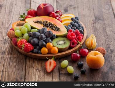 Fresh raw organic summer berries and exotic fruits in round wooden plate on light wooden kitchen background. Papaya, grapes, nectarine, orange, raspberry, kiwi, strawberry, lychees, cherry. Top view
