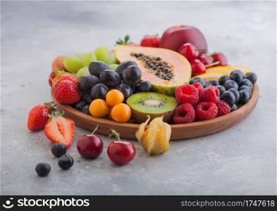 Fresh raw organic summer berries and exotic fruits in round wooden plate on light kitchen background. Papaya, grapes, nectarine, orange, raspberry, kiwi, strawberry, lychees, cherry. Top view