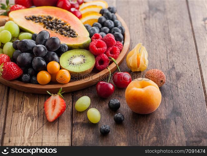 Fresh raw organic summer berries and exotic fruits in round wooden plate on wooden kitchen background. Papaya, grapes, nectarine, orange, raspberry, kiwi, strawberry, lychees, cherry Top view