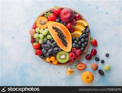 Fresh raw organic summer berries and exotic fruits in round wooden plate on blue kitchen background. Papaya, grapes, nectarine, orange, raspberry, kiwi, strawberry, lychees, cherry. Top view