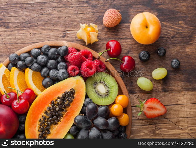 Fresh raw organic summer berries and exotic fruits in round wooden plate on wooden kitchen background. Papaya, grapes, nectarine, orange, raspberry, kiwi, strawberry, lychees, cherry.Top view