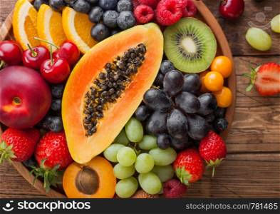 Fresh raw organic summer berries and exotic fruits in round wooden plate on wooden kitchen background. Papaya, grapes, nectarine, orange, raspberry, kiwi, strawberry, lychees, cherry.Top view
