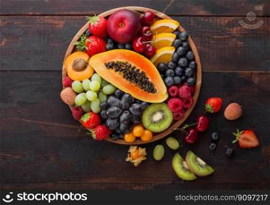 Fresh raw organic summer berries and exotic fruits in round wooden plate on dark wooden kitchen background. Papaya, grapes, nectarine, orange, raspberry, kiwi, strawberry, lychees, cherry. Top view
