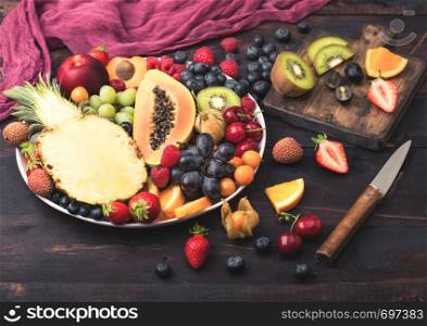 Fresh raw organic summer berries and exotic fruits in white plate on dark wooden background with chopping board and knife. Pineapple, papaya, grapes, nectarine, orange, apricot, kiwi, pear. Top view