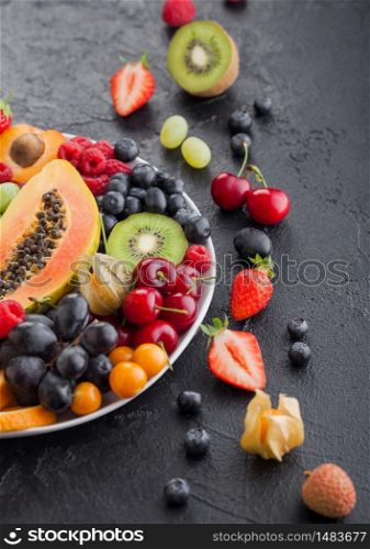Fresh raw organic summer berries and exotic fruits in white plate on black background. Pineapple, papaya, grapes, nectarine, orange, apricot, kiwi, pear, lychees, cherry and physalis. Top view