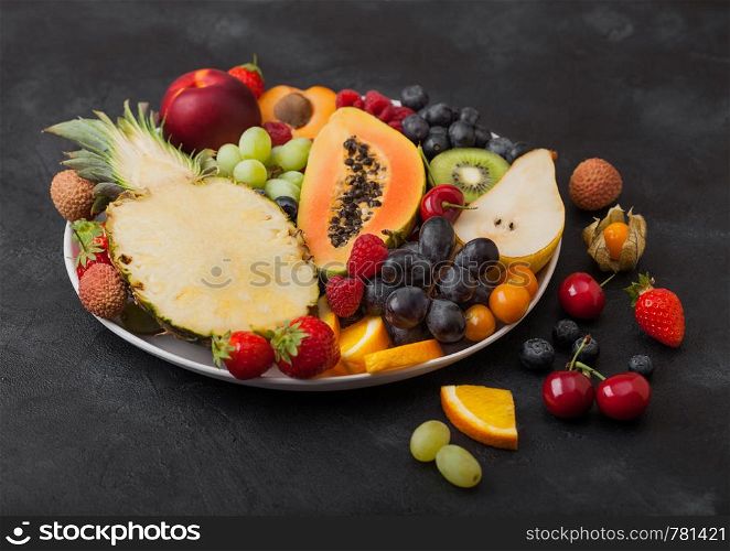 Fresh raw organic summer berries and exotic fruits in white plate on black background. Pineapple, papaya, grapes, nectarine, orange, apricot, kiwi, pear, lychees, cherry and physalis. Top view
