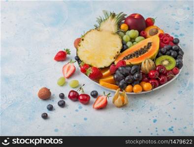 Fresh raw organic summer berries and exotic fruits in white plate on blue kitchen background. Pineapple, papaya, grapes, nectarine, orange, apricot, kiwi, pear, lychees, cherry and physalis. Top view
