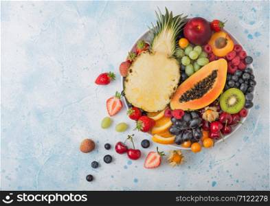 Fresh raw organic summer berries and exotic fruits in white plate on blue kitchen background. Pineapple, papaya, grapes, nectarine, orange, apricot, kiwi, pear, lychees, cherry and physalis. Top view