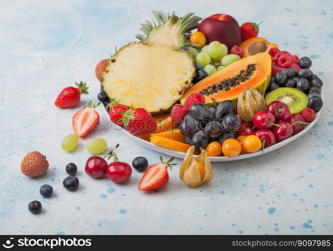 Fresh raw organic summer berries and exotic fruits in white plate on blue kitchen background. Pineapple, papaya, grapes, nectarine, orange, apricot, kiwi, pear, lychees, cherry and physalis. Top view