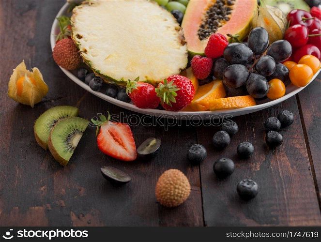 Fresh raw organic summer berries and exotic fruits in white plate on dark wooden background. Pineapple, papaya, grapes, nectarine, orange, apricot, kiwi, strawberry, lychees, cherry. Top view