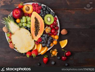 Fresh raw organic summer berries and exotic fruits in white plate on dark wooden background. Pineapple, papaya, grapes, nectarine, orange, apricot, kiwi, strawberry, lychees, cherry. Top view