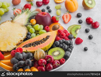 Fresh raw organic summer berries and exotic fruits in white plate on light background. Pineapple, papaya, grapes, nectarine, orange, apricot, kiwi, pear, lychees, cherry and physalis. Top view