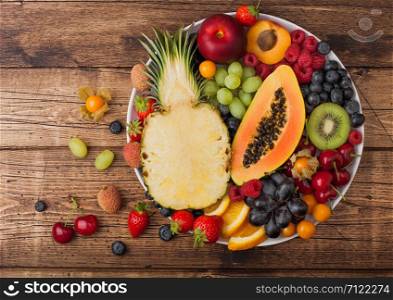 Fresh raw organic summer berries and exotic fruits in white plate on vintage wooden background. Pineapple, papaya, grapes, nectarine, orange, apricot, kiwi, pear, lychees, cherry and physalis. Top view