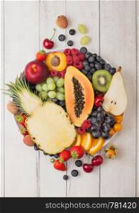 Fresh raw organic summer berries and exotic fruits in white plate on white wooden background. Pineapple, papaya, grapes, nectarine, orange, apricot, kiwi, pear, lychees, cherry and physalis. Top view