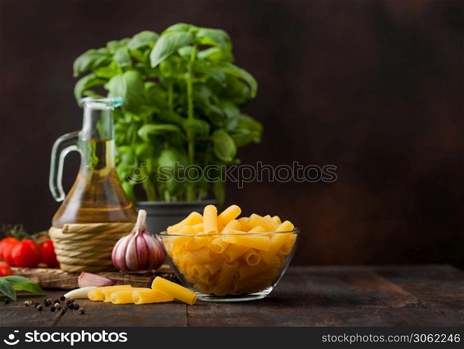 Fresh raw penne pasta in glass bowl with basil plant, oil and tomatoes with garlic and pepper on wooden table background.