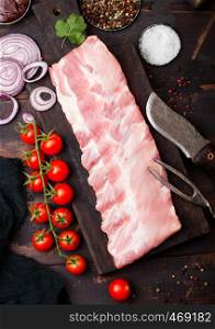 Fresh raw pork ribs on chopping board and vintage meat fork and knife on wooden background. Fresh tomatoes and red onion with garlic, salt and pepper and kitchen towel.