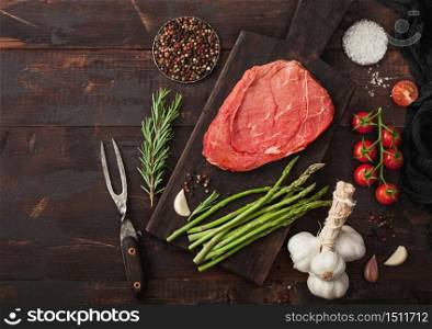 Fresh raw slice of beef braising steak on chopping board with asparagus, garlic and tomatoes with salt and pepper on wooden table background.