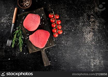 Fresh raw tuna on a cutting board with tomatoes, spices and rosemary. On a black background. High quality photo. Fresh raw tuna on a cutting board with tomatoes, spices and rosemary.