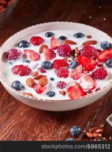 Fresh ripe berries, almonds, nuts, oatmeal and granola in a plate with milk on a wooden background with copy space. Healthy dietary food.. Dietary homemade natural breakfast with fresh organic ingredients - berries, granola, nuts and milk in a white bowl on a wooden table.