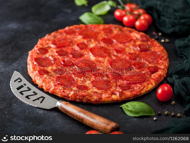 Fresh round baked Hot and Spicy Pepperoni pizza with knife and tomatoes with basil on black kitchen table background.
